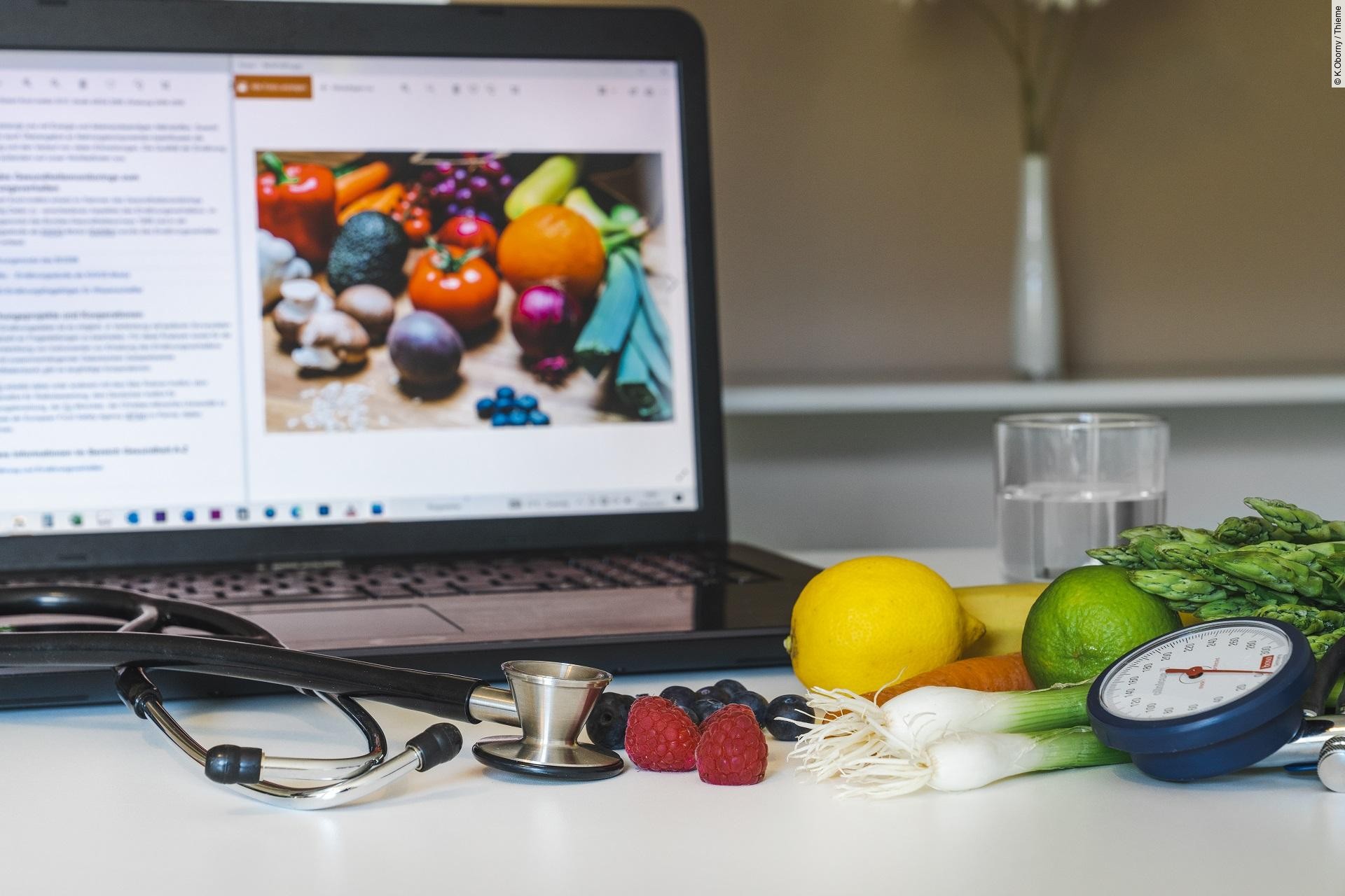 Laptop, vor dem ein Glas Wasser, Stethoskop, Blutdruckmessgerät, Obst und Gemüse sind.