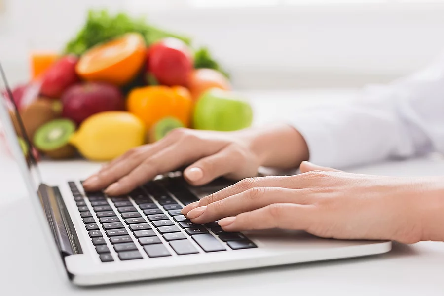 Ernährungsberaterin tippt auf Tastatur, daneben liegen Obst und Gemüse
