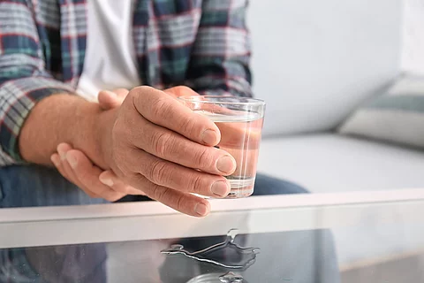 Mann hält seinen rechten Arm mit der linken Hand fest, in der rechten Hand hat er ein Glas Wasser.