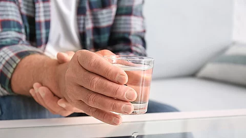 Mann hält seinen rechten Arm mit der linken Hand fest, in der rechten Hand hat er ein Glas Wasser.