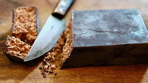 Ein Stück Rokakao, abgeschnitten von einem Block aus Rohkakao auf einem dunkelbraunen Holztisch mit einem Messer. 