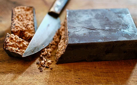 Ein Stück Rokakao, abgeschnitten von einem Block aus Rohkakao auf einem dunkelbraunen Holztisch mit einem Messer. 
