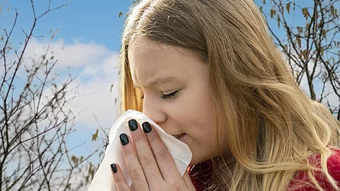 Frau niest ins Taschentuch, Heuschnupfenzeit, im Hintergrund Baumpollen