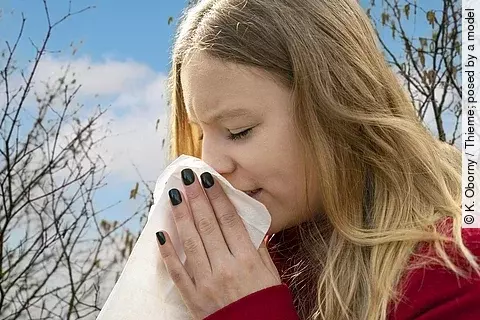 Frau niest ins Taschentuch, Heuschnupfenzeit, im Hintergrund Baumpollen