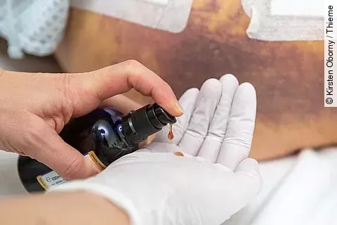 Hand in weißem Handschuh mit Aromaöl Flasche. Im Hintergrund ist ein Oberschenkel mit einem Hämatom zu sehen. 