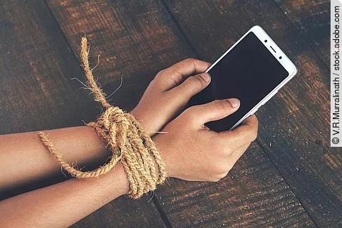 Gefesselte Kinderhände mit Smartphone in der Hand; Symbolbild für Handysucht