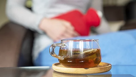 Frau mit Wärmflasche auf dem Unterleib, Glas Tee auf dem Tisch davor