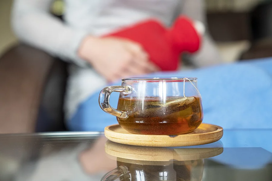 Frau mit Wärmflasche auf dem Unterleib, Glas Tee auf dem Tisch davor