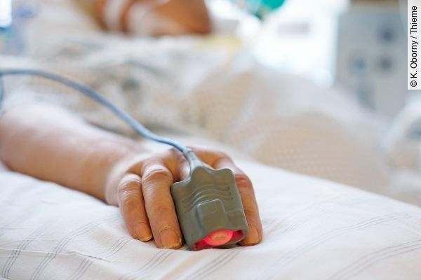 Patient liegt in seinem Bett auf der Intensivstation. Person ist nicht erkennbar, nur die Hand mit einem Finger im Pulsoximeter. 