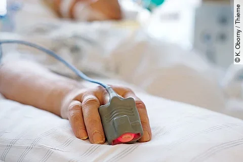 Patient liegt in seinem Bett auf der Intensivstation. Person ist nicht erkennbar, nur die Hand mit einem Finger im Pulsoximeter. 