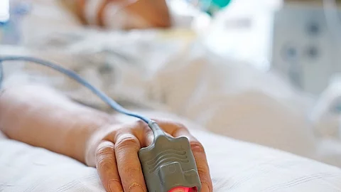 Patient liegt in seinem Bett auf der Intensivstation. Person ist nicht erkennbar, nur die Hand mit einem Finger im Pulsoximeter. 