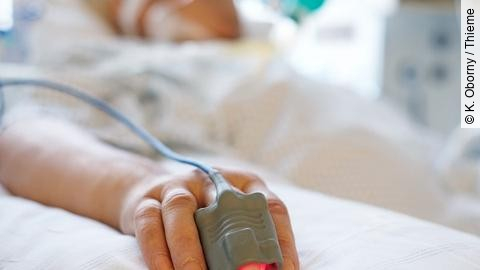 Patient liegt in seinem Bett auf der Intensivstation. Person ist nicht erkennbar, nur die Hand mit einem Finger im Pulsoximeter. 