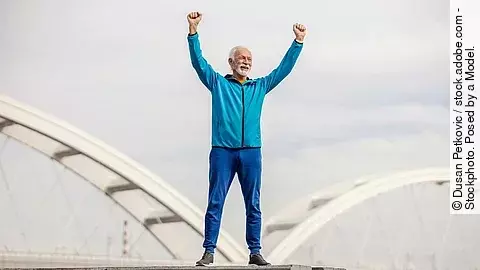 Senior in Sportkleidung mit nach oben gestreckten Armen vor einer Brücke