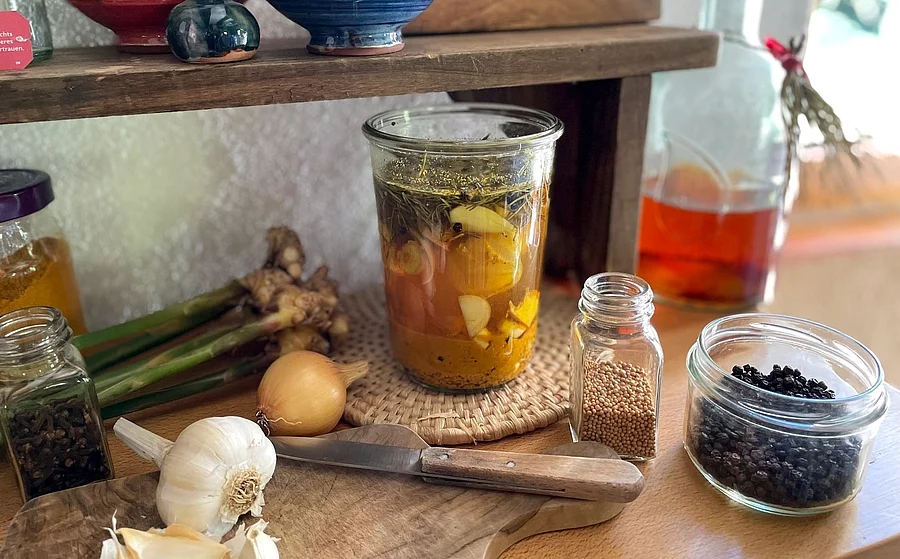 Zutaten für Fire Cider auf Küchenarbeitsplatte: Knoblauch, Weckglas mit Apfelessig, Pfefferkörner, Zwiebel, Holzbrett mit Messer