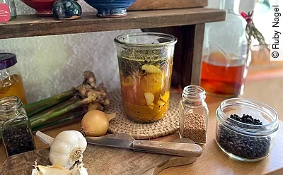 Zutaten für Fire Cider auf Küchenarbeitsplatte: Knoblauch, Weckglas mit Apfelessig, Pfefferkörner, Zwiebel, Holzbrett mit Messer