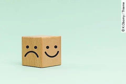 Holzwürfel mit einem lachenden und traurigen Smiley auf türkisem Hintergrund.