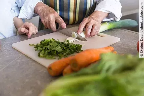 Nahaufnahme einer älteren Dame, die Gemüse auf einem Brett schneidet.