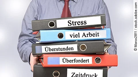 Arbeitnehmer hält Ordner mit den Aufschriften Stress, viel Arbeit, Überstunden, Überfordert und Zeitdruck
