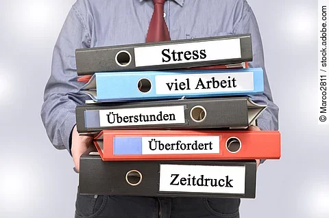 Arbeitnehmer hält Ordner mit den Aufschriften Stress, viel Arbeit, Überstunden, Überfordert und Zeitdruck