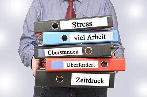 Arbeitnehmer hält Ordner mit den Aufschriften Stress, viel Arbeit, Überstunden, Überfordert und Zeitdruck
