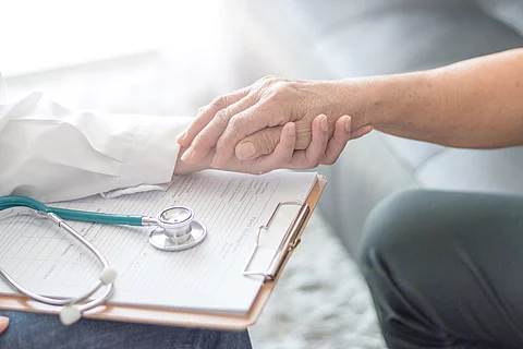 Ärztin mit Klemmbrett und Stethoskop hält die Hand eines Patientens.
