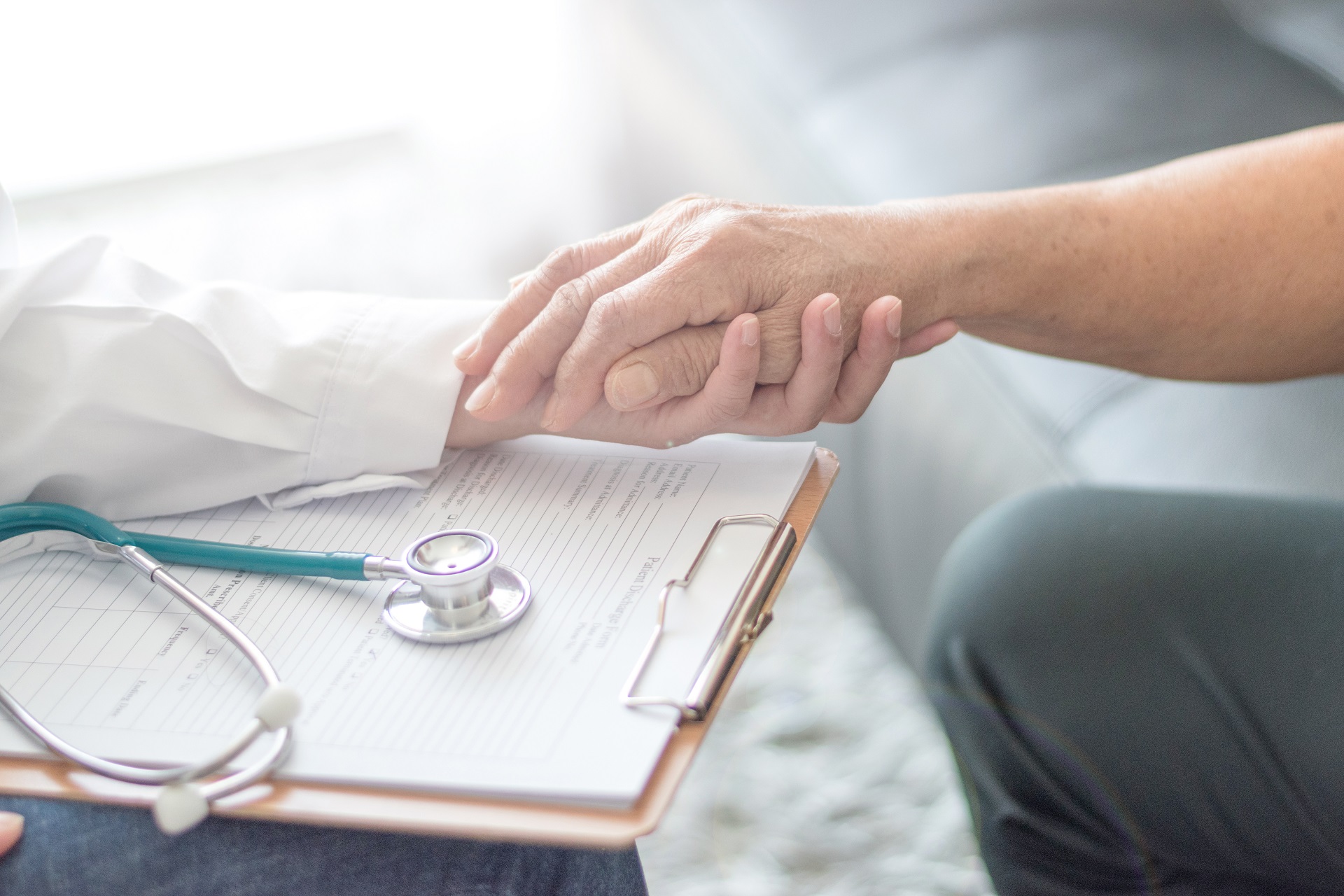 Ärztin mit Klemmbrett und Stethoskop hält die Hand eines Patientens.