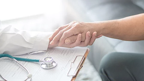 Ärztin mit Klemmbrett und Stethoskop hält die Hand eines Patientens.