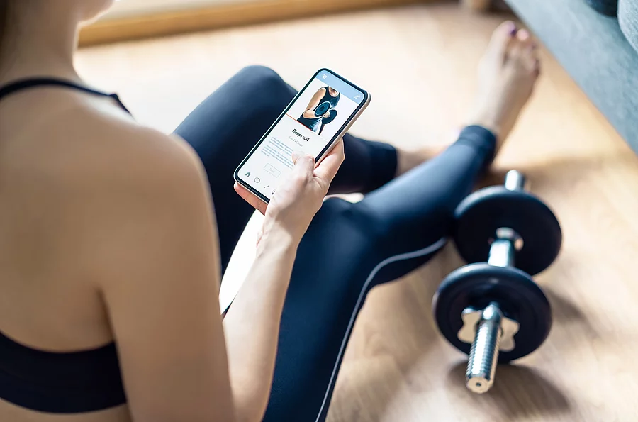 Frau in Sportsachen sitzt mit Trainingsanleitung auf dem Smartphone auf dem Boden, neben ihr liegen Hanteln