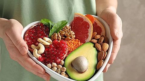 Person hält eine herzförmige Schüssel mit Obst, Hülsenfrüchten und Nüssen hoch.