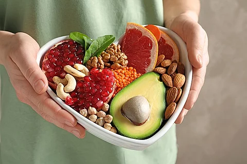 Person hält eine herzförmige Schüssel mit Obst, Hülsenfrüchten und Nüssen hoch.