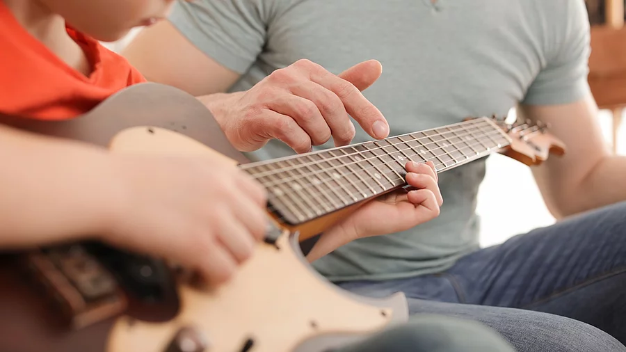 Kind spielt Gitarre, Erwachsener daneben leitet ihn an Kind spielt Gitarre, Erwachsener daneben leitet ihn an