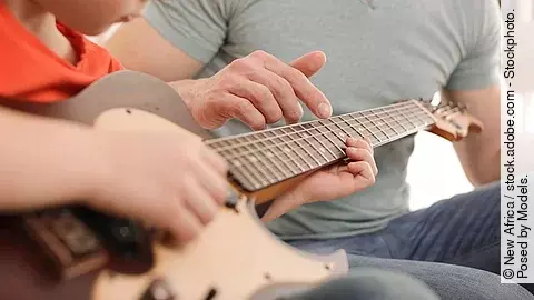 Kind spielt Gitarre, Erwachsener daneben leitet ihn an