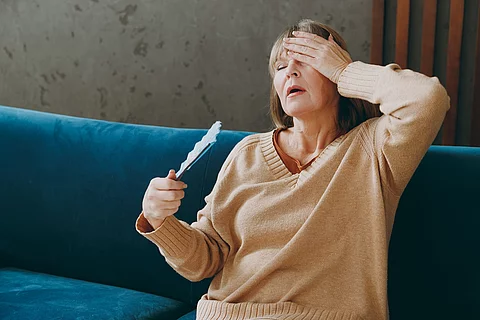 Ältere Frau mit Hitzewallung auf dem Sofa, fächert sich Luft zu