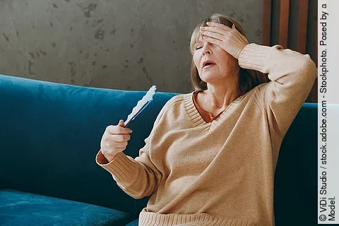 Ältere Frau mit Hitzewallung auf dem Sofa, fächert sich Luft zu