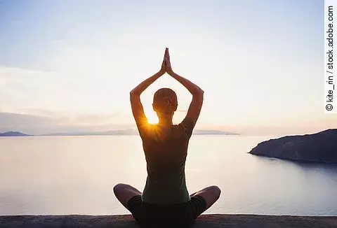 Frau sitzt am Seeufer und macht Yoga.