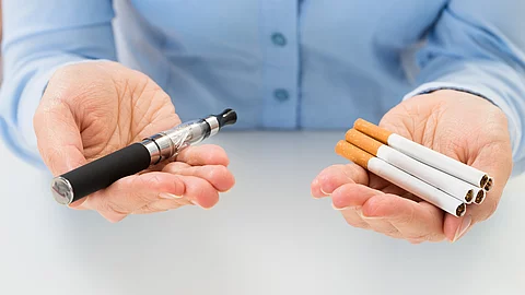 Eine Frau mit blauer Bluse hält in der linken Hand einige Zigaretten und in der rechten Hand eine E-Zigarette. 