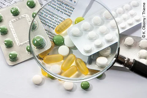 Durch eine Lupe sind verschiedene Nahrungsergänzungsmittel zu sehen. Zum Teil noch im Bilster, zum Teil als freie Tabletten auf dem Tisch. 