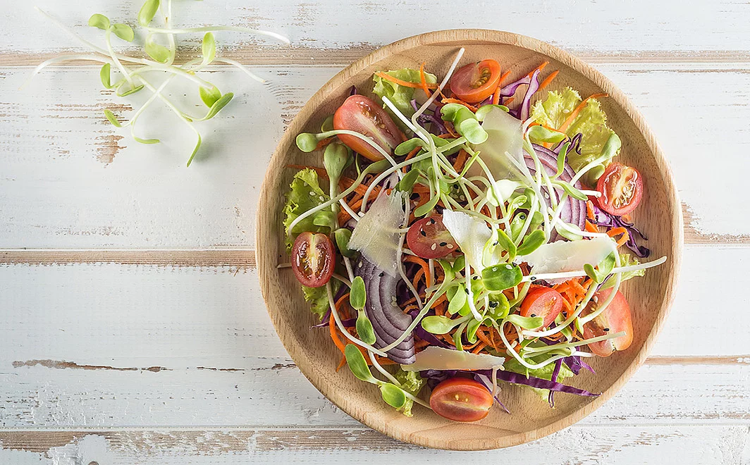 Holzschale gefüllt mit Salat aus Microgreens von Sonnenblumen, Tomaten, Karotten, Zwiebeln und Sesam. Daneben liegen ein paar Microgreens von Sonnenblumen.