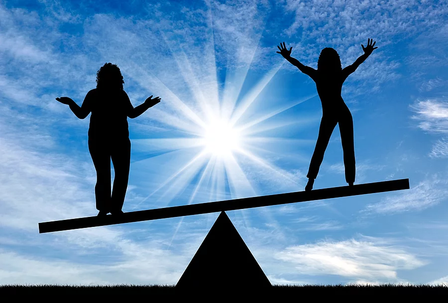 2 Frauen stehen auf einer Wippe, rechts ohne, links mit Übergewicht