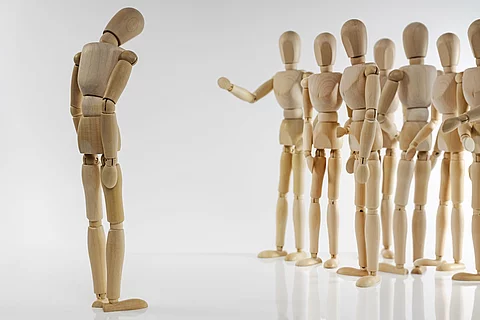 eine Holzpuppe mit gesenktem Kopf steht einer Gruppe von Holzpuppen gegenüber