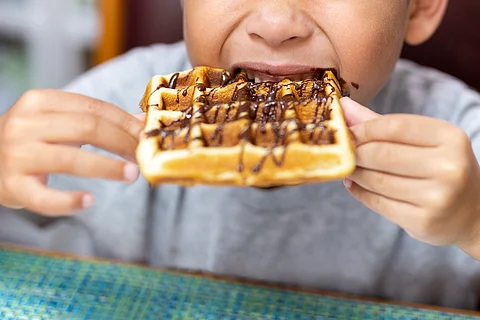 Child bitting waffle with chocolate syrup at breakfast table.