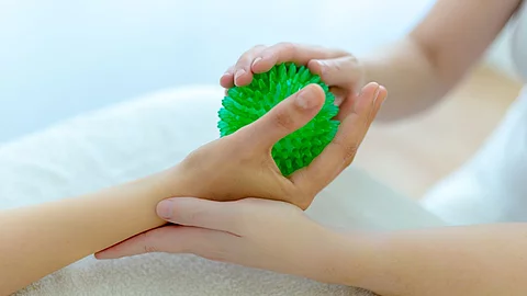Eine Person hält einen grünen Igelball in der Hand, während eine weitere Person sie dabei mit beiden Händen unterstützt.