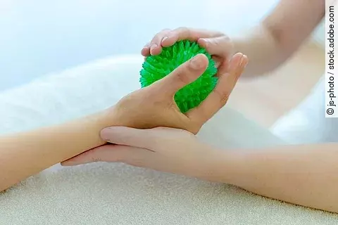 Eine Person hält einen grünen Igelball in der Hand, während eine weitere Person sie dabei mit beiden Händen unterstützt.