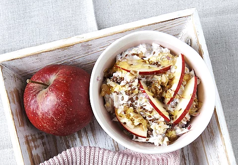 Birchermüsli auf weißem Tablett, daneben ein Apfel