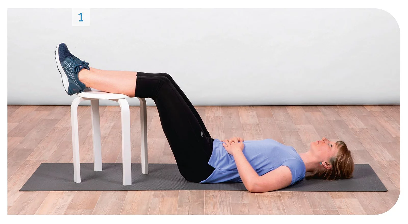 Junge Frau in Sportkleidung liegt auf Yogamatte auf dem Rücken und hat die Oberschenkel auf einem Hocker abgelegt.