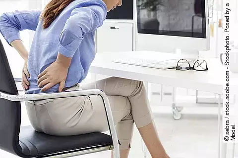 Frau sitzt am Schreibtisch und hält sich ihren schmerzenden Rücken