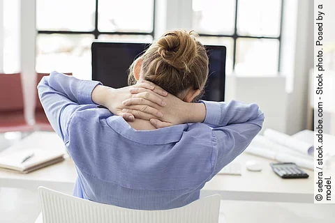 Eine Frau sitzt am Schreibtisch und stützt mit den Händen den Nacken.
