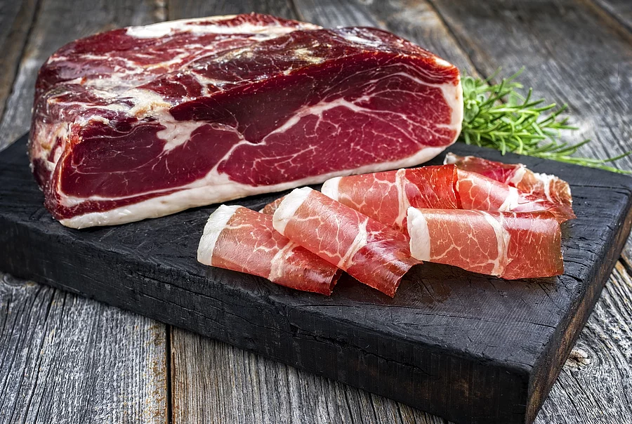 Traditioneller luftgetrockneter Parma Schinken mit Kräutern angeboten als closeup auf einem rustikalen verbrannten Schneidebrett Schinken auf grauem Brett