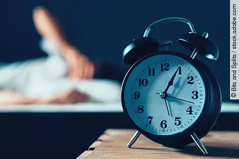 Im Vordergrund steht ein Wecker, im Hintergrund liegt eine Person im Bett.