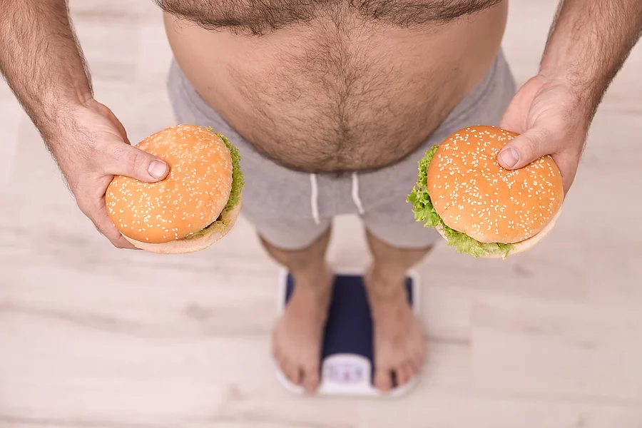Overweight man holding burgers while measuring his weight using Übergewichtiger auf der Waage, in beiden Händen ein Burger